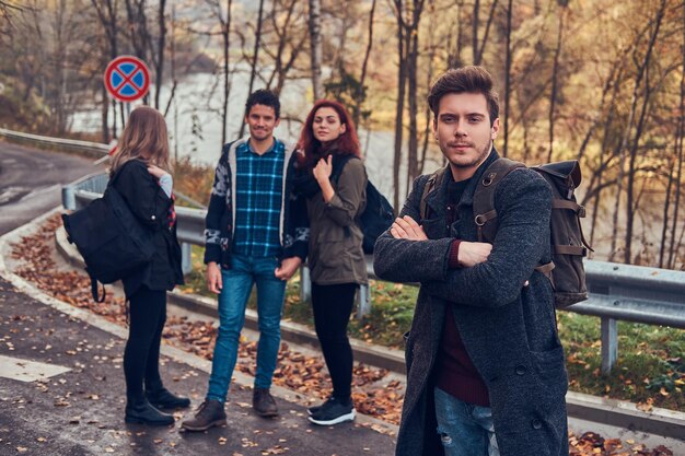 Viaggio, autostop, concetto di avventura. Gruppo di giovani escursionisti in piedi ai margini della strada nella bellissima foresta autunnale.