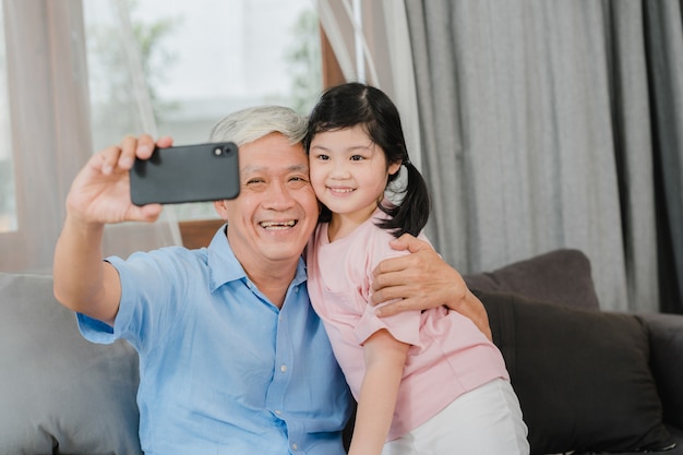 Videochiamata asiatica della nipote e del nonno a casa. Nonno cinese senior soddisfatto della ragazza che utilizza la videochiamata del telefono cellulare che parla con suo papà e mamma che si trovano nel salone a casa.