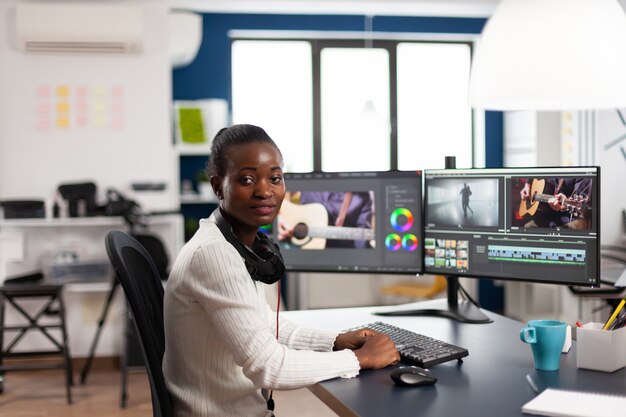 Videografo nero che sorride alla telecamera che modifica il progetto video nel software di post produzione che lavora nell'ufficio dello studio creativo