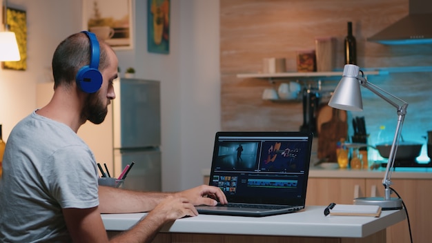 Videografo professionista che lavora nell'app di editing video indossando le cuffie davanti al laptop seduto nella cucina di casa. Freelancer elabora il montaggio di film audio su laptop professionale a mezzanotte