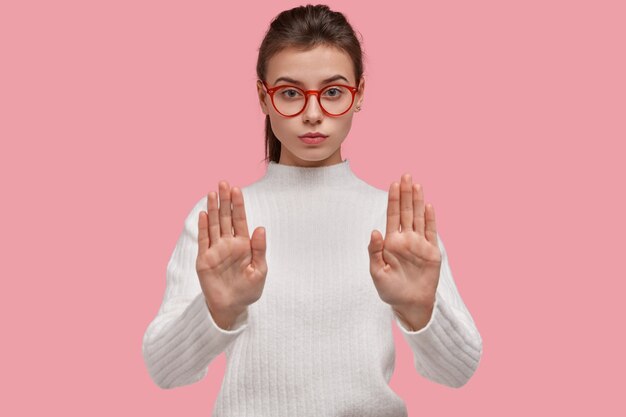 Vietato oltrepassare. La femmina attraente seria tira le mani verso la macchina fotografica, mostra il gesto di arresto, rifiuta qualcosa, indossa gli occhiali