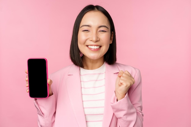 Vincere donna d'affari coreana che mostra lo schermo dello smartphone sorridente soddisfatto dimostrando il negozio online di applicazioni per telefoni cellulari o l'app per lo shopping in piedi su sfondo rosa