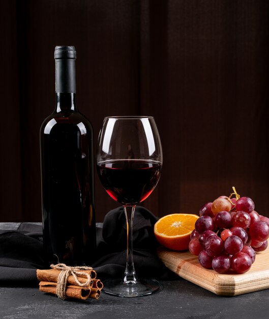Vino rosso di vista laterale con l'uva, arancia sul tagliere di legno sul verticale scuro