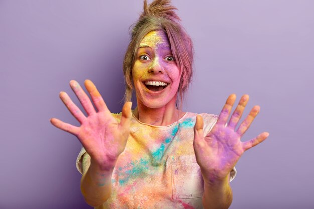 Visita il festival dei colori di Holi. La donna sorridente felice ha spruzzi colorati su se stessa, sporca di polvere, mostra palme dipinte multicolori, isolate su un muro viola. Concetto di celebrazione