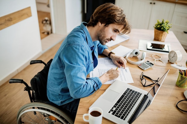 Vista ad alto angolo di giovane uomo d'affari in sedia a rotelle che fa scartoffie mentre lavora al computer portatile a casa