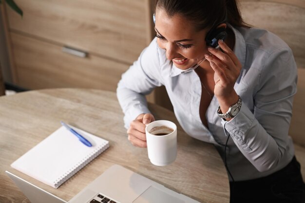 Vista ad alto angolo di un felice agente del call center che beve caffè mentre comunica con un cliente al telefono