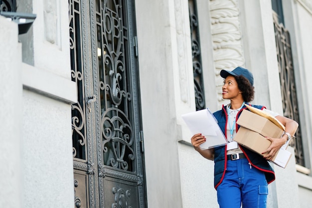 Vista ad angolo basso del sorridente lavoratore postale afroamericano che trasporta pacchi mentre effettua la consegna nel quartiere residenziale