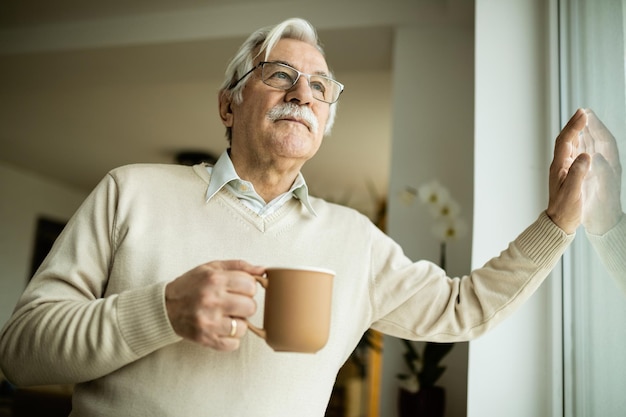 Vista ad angolo basso dell'uomo anziano che sogna ad occhi aperti mentre beve caffè e guarda attraverso la finestra