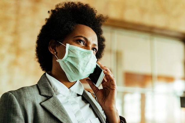 Vista ad angolo basso della donna d'affari nera che indossa una maschera protettiva durante l'epidemia di virus e comunica sul telefono cellulare mentre si trova nel corridoio dell'aeroporto