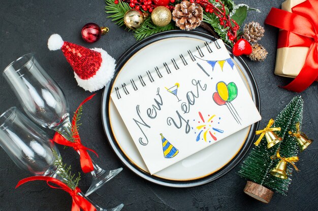 Vista aerea del taccuino a spirale con la penna sul piatto di cena albero di natale rami di abete cono di conifera confezione regalo cappello di babbo natale calici di vetro caduti su sfondo nero