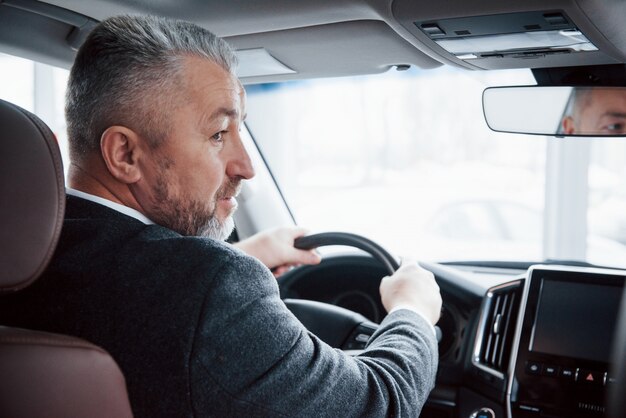 Vista da dietro dell'uomo d'affari maggiore in abiti ufficiali alla guida di una nuova auto moderna