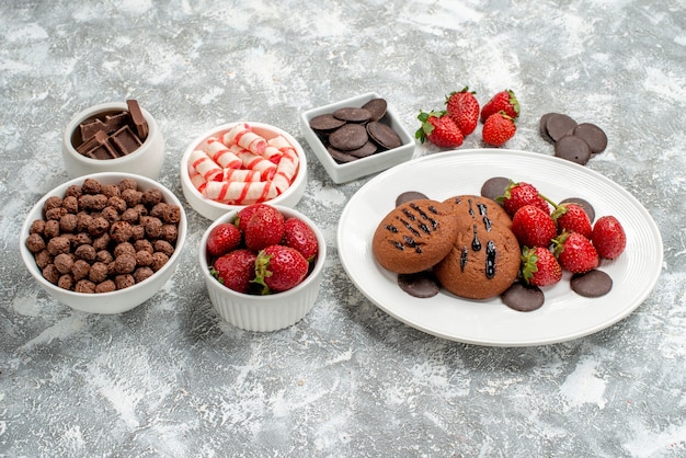Vista dal basso biscotti fragole e cioccolatini rotondi sulla piastra ovale ciotole con caramelle fragole cioccolatini cereali sul tavolo grigio-bianco