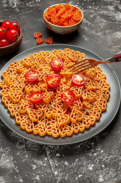 Vista dal basso cuori di pasta italiana tagliati pomodorini su piastra ovale forchetta cuore rosso pasta in ciotola sul tavolo grigio