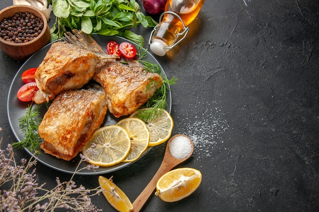 Vista dal basso gustosa frittura di pesce fette di limone tagliate pomodorini su piastra forchetta e coltello bottiglia di olio menta sul tavolo nero copia posto