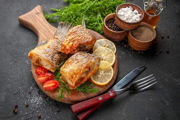 Vista dal basso gustosa frittura di pesce fette di limone tagliate pomodorini sul tagliere spezie diverse in ciotole coltello e forchetta su sfondo nero