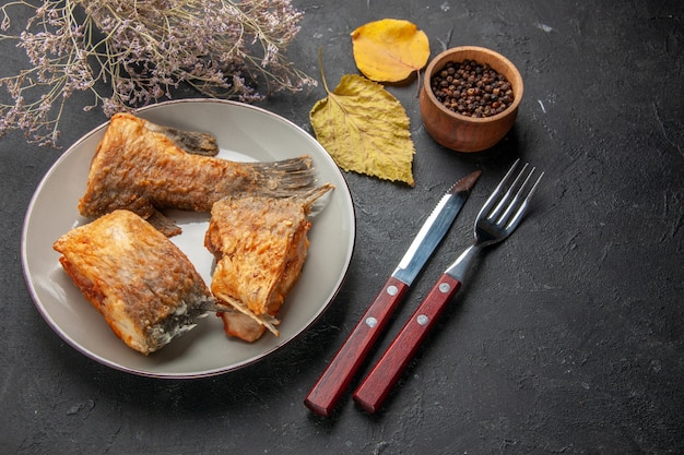 Vista dal basso gustosa frittura di pesce su forchetta e coltello ramo di fiori secchi pepe nero in ciotola di legno su tavola nera