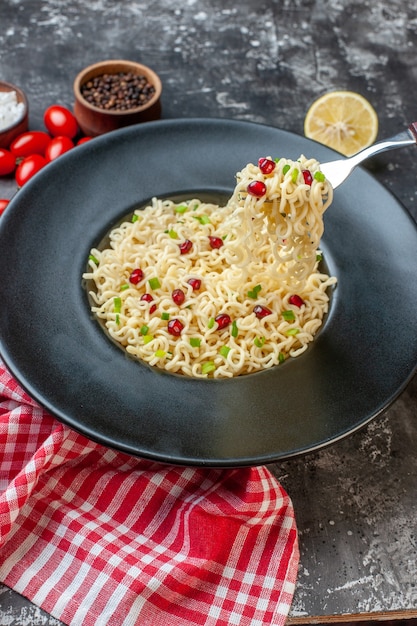 Vista dal basso ramen noodles sollevati su una forchetta su piastra nera pepe nero in una ciotola pomodorini tagliati al limone sul tavolo scuro