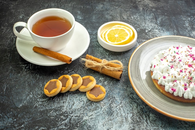 Vista dal basso tazza di tè aromatizzata da fette di limone alla cannella in piccoli biscotti con piattino bastoncini di cannella sul tavolo scuro