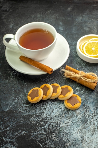 Vista dal basso tazza di tè aromatizzata da fette di limone alla cannella in piccoli biscotti con piattino bastoncini di cannella sul tavolo scuro
