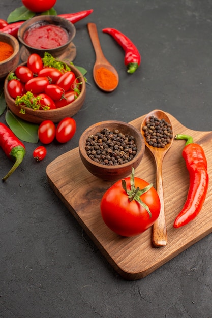 Vista dal basso una ciotola di pomodorini peperoni rossi piccanti foglie di alloro e una ciotola di pepe nero un cucchiaio di legno un pomodoro un peperone rosso sul tagliere su sfondo nero