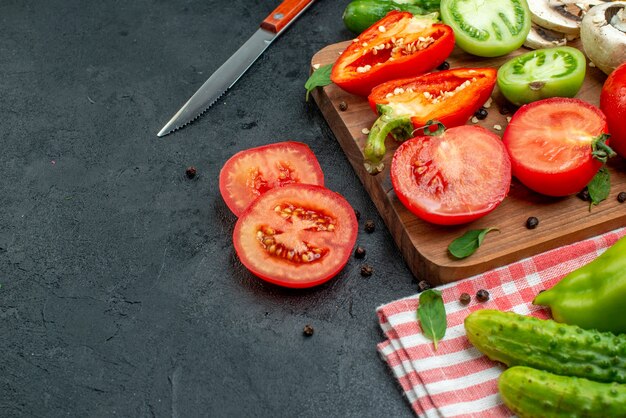 Vista dal basso verdure pomodori peperoni sul tagliere verdure in una ciotola cetrioli su tovaglia rossa coltello sul tavolo nero copia posto