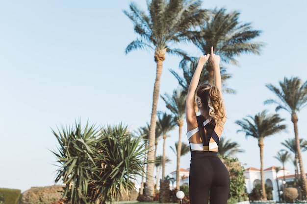 Vista dal retro gioiosa giovane donna in abiti sportivi che si estende su palme e cielo blu. Mattina soleggiata, espressione di positività, vere emozioni, stile di vita sano.