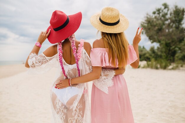 Vista dal retro su due belle donne alla moda in spiaggia in vacanza, stile estivo, tendenza della moda, cappelli di paglia con indosso, tendenza della moda, abito rosa e pizzo, vestito sexy