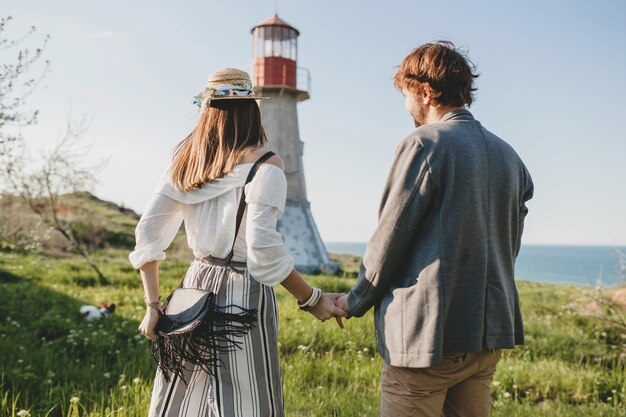 Vista dal retro su stile indie giovane coppia hipster innamorato camminando in campagna, tenendosi per mano, faro sullo sfondo, calda giornata estiva, soleggiato, vestito bohémien, cappello