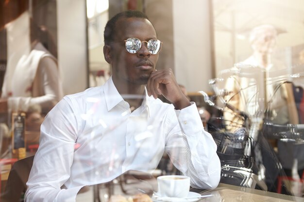 Vista dal vetro della finestra del bel CEO afroamericano pensieroso in camicia bianca e tonalità eleganti con caffè nero durante il pranzo, seduto da solo al caffè, con uno sguardo pensieroso, toccando il mento