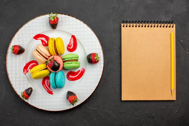 Foto gratuito vista dall'alto amaretto francese fragole ricoperte di cioccolato e amaretti francesi accanto al quaderno crema e matita gialla sul tavolo scuro