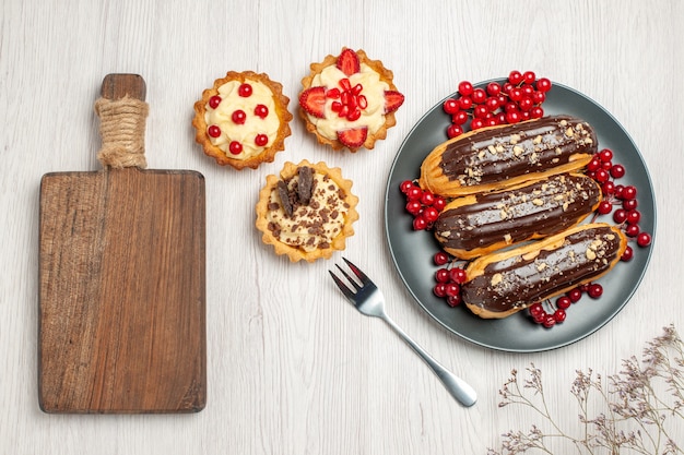 Vista dall'alto bignè al cioccolato e ribes sui biscotti della piastra grigia, una forchetta e un tagliere sul tavolo di legno bianco