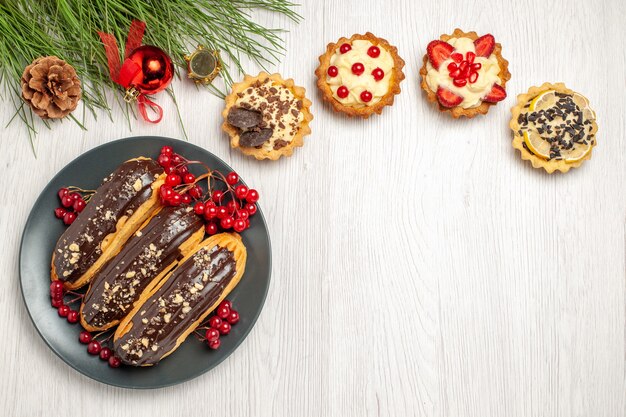Vista dall'alto bignè al cioccolato e ribes sulle torte della banda grigia e foglie di pino con i giocattoli di Natale sul tavolo di legno bianco