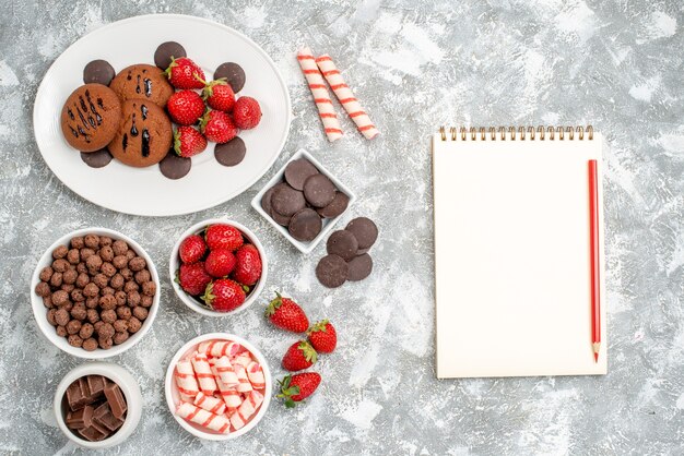 Vista dall'alto biscotti fragole e cioccolatini rotondi sul piatto ovale ciotole con caramelle fragole cioccolatini cereali e un taccuino con matita sul tavolo grigio-bianco