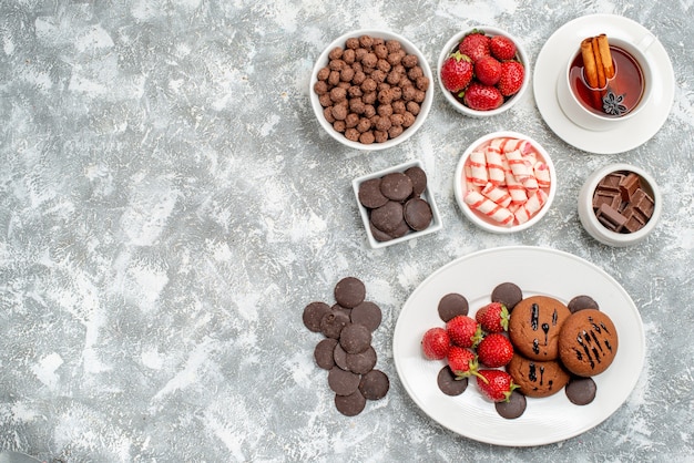 Vista dall'alto biscotti fragole e cioccolatini rotondi sul piatto ovale ciotole con caramelle fragole e cioccolatini cereali e una tazza di tè sul lato destro del tavolo grigio-bianco con spazio libero