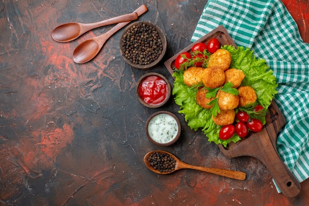 Vista dall'alto bocconcini di pollo lattuga pomodorini su tavola di legno pepe nero in ciotola salse in piccole ciotole di legno cucchiai di legno su tavolo scuro spazio libero