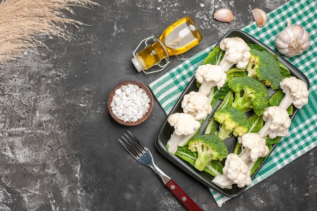 Vista dall'alto broccoli crudi e cavolfiore su piastra rettangolare nera su tovagliolo a scacchi verde e bianco forchetta olio aglio sale marino su superficie scura spazio libero