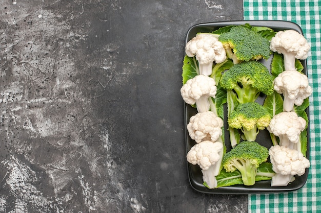 Vista dall'alto broccoli crudi e cavolfiore sulla piastra rettangolare nera sul tovagliolo a scacchi verde e bianco su superficie scura con spazio di copia