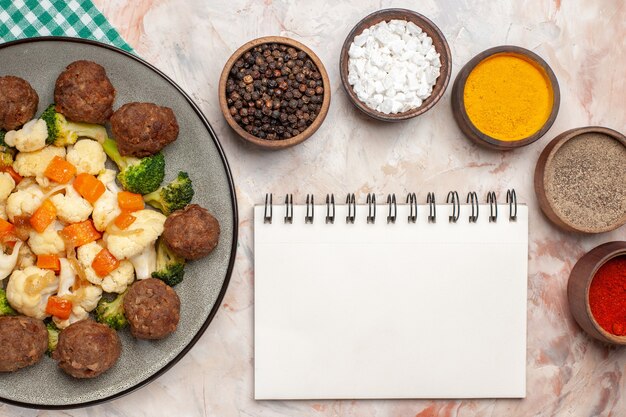 Vista dall'alto broccoli e cavolfiore insalata e polpetta su piatto tovagliolo a scacchi verde e bianco spezie colorate in piccole ciotole un blocco note su sfondo nudo isolato