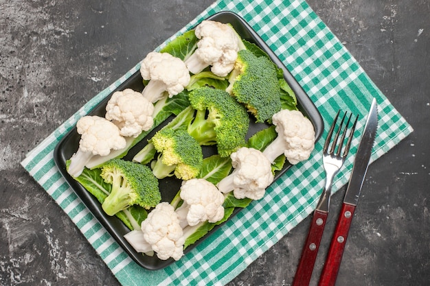 Vista dall'alto broccoli e cavolfiore su piatto rettangolare nero su forchetta e coltello per tovaglioli a scacchi verdi e bianchi su sfondo scuro