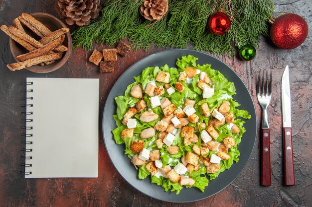 Vista dall'alto Caesar Salad su piastra ovale forchetta coltello ciotola con crosta secca albero di Natale giocattoli un quaderno su sfondo rosso scuro