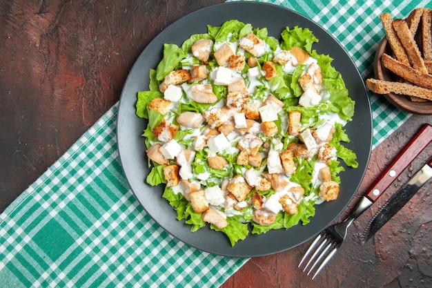 Vista dall'alto Caesar Salad su piastra ovale tovaglia a scacchi bianchi verdi forchetta e coltello su sfondo rosso scuro