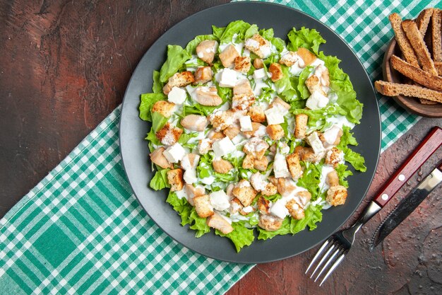 Vista dall'alto Caesar Salad su piastra ovale tovaglia a scacchi bianchi verdi forchetta e coltello su sfondo rosso scuro