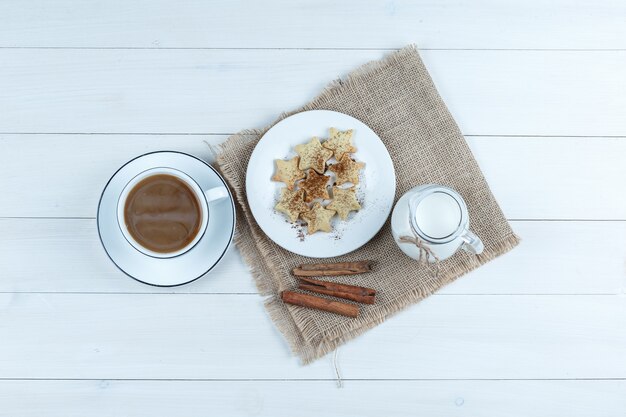 Vista dall'alto caffè in tazza con biscotti, bastoncini di cannella, latte su fondo di legno e pezzo di sacco.