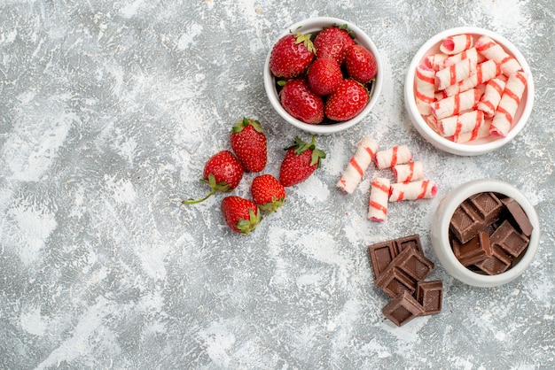 Vista dall'alto ciotole con fragole cioccolatini caramelle e alcune fragole cioccolatini caramelle sul lato destro del mosaico grigio-bianco di massa