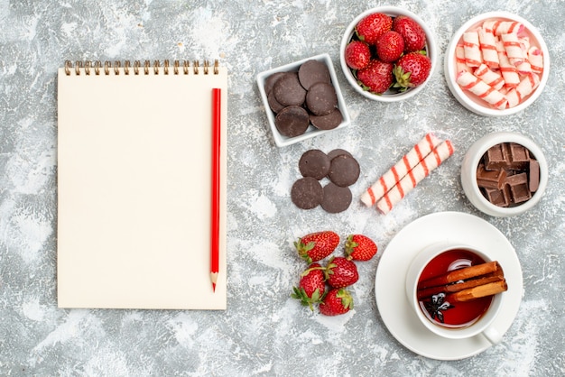 Vista dall'alto ciotole con fragole cioccolatini caramelle e tè ai semi di anice cannella e taccuino con matita sul fondo grigio-bianco