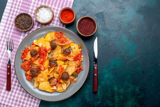 Vista dall'alto cotte patate a fette con polpette di carne insieme a condimenti sulla scrivania blu scuro.