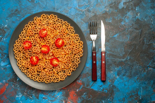 Vista dall'alto cuori di pasta italiana tagliati pomodorini su forchetta e coltello piatto ovale nero su spazio libero tavolo blu