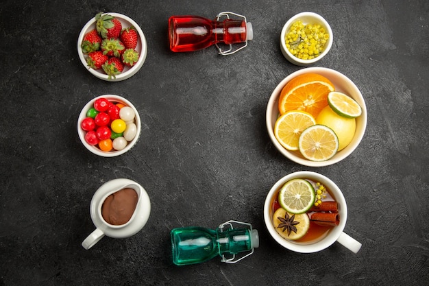 Vista dall'alto da lontano bacche e ciotole di cioccolato di crema al cioccolato caramelle erbe fragole e limoni a fette accanto alla tazza di tè e bottiglie colorate