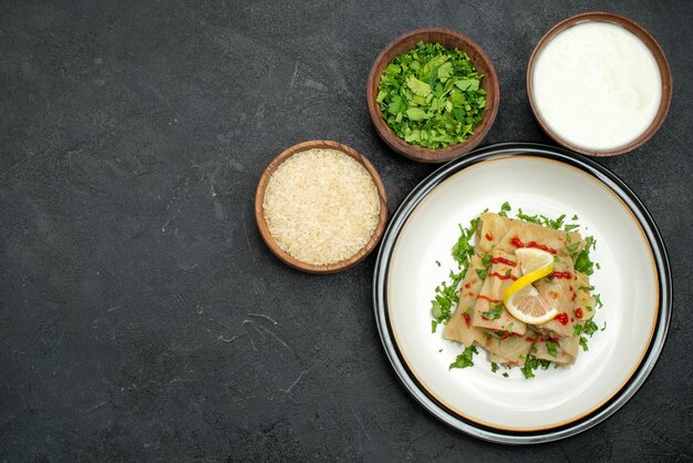 Vista dall'alto da lontano cavolo ripieno appetitoso cavolo ripieno con erbe limone e salsa su piatto bianco e ciotole con erbe di riso e panna acida sul lato destro del tavolo nero