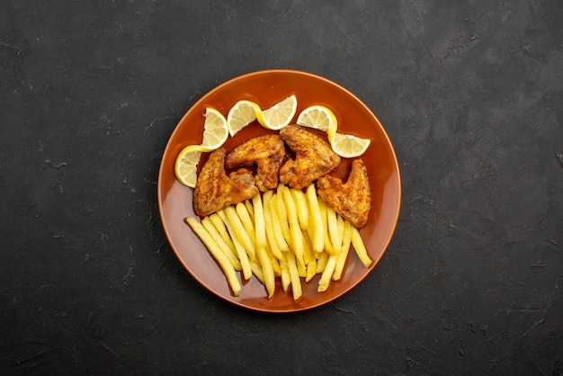 Vista dall'alto da lontano cibo su ali di pollo piatto con patatine fritte e limone su piatto arancione sul tavolo scuro